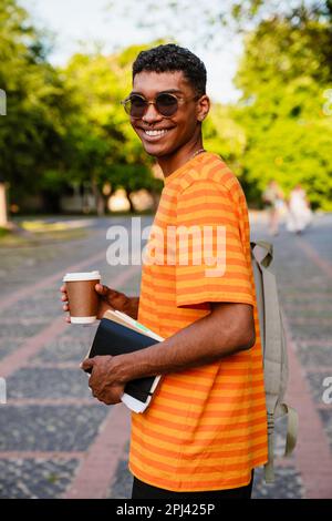 Giovane allievo allegro dell'uomo africano in vestito casuale che tiene il caffè ed i libri mentre si levano in piedi all'aperto Foto Stock