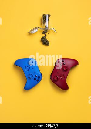 Due gamepad wireless, rosso e blu, giacciono su uno sfondo giallo, accanto a un vaso aperto con boccioli di marijuana secchi. Vacanze a casa, weekend di divertimento, happines Foto Stock