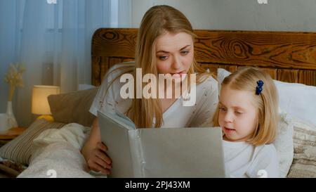 Concetto di apprendimento familiare. Madre caucasica aiutare piccolo carino figlia prescolare bambino ragazza libro lettura libro a letto a casa mamma camera da letto con bambino Foto Stock