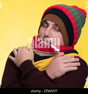 Un giovane uomo in abiti caldi su sfondo giallo tiene in bocca un termometro elettronico ed è sconvolto dall'alta temperatura Foto Stock