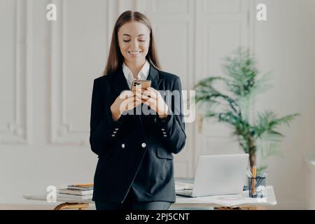 Felice professionista imprenditore femminile in tuta formale che tiene il telefono e chattare con un amico o leggere notizie positive, donna d'affari sorridente e looki Foto Stock