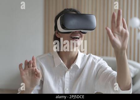 Una libera donna felice che indossa un visore VR mentre lavora in remoto online da casa, sollevando le mani cercando di toccare gli oggetti in realtà virtuale. Ragazza Foto Stock