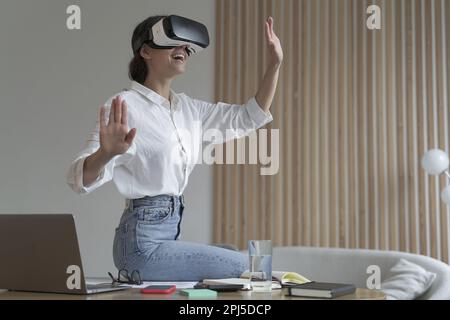 Tecnologie future. Giovane donna d'affari stupito in occhiali di realtà virtuale che testano metodo innovativo per il business, seduto sulla scrivania al suo posto di lavoro e Foto Stock