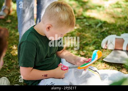 Svetlogorsk, Russia - 13.08.2022 - il ragazzino taglia un pezzo di carta con le forbici alla lezione di arte all'aperto di carta artigianale, insegnando ai bambini a creare carta c Foto Stock