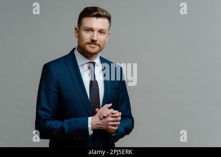 Stile formale. Serio imprenditore maschile bearded tiene le mani insieme indossa abiti formali vestiti vestiti in ufficio appese contro sfondo grigio con Foto Stock