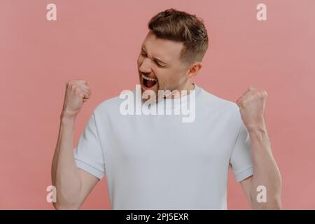 Studio ritratto di felice uomo overjoyed in bianco tshirt bianca godendo la vittoria o il successo, ragazzo eccitato pugni clenching e facendo sì gesto mentre sta Foto Stock