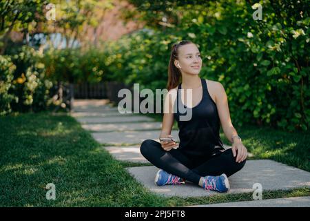 La giovane donna europea dai capelli scuri siede incroci le gambe guarda da parte con l'espressione soddisfatta usa smartphone ha una pausa dopo l'allenamento cardio vestito in ac Foto Stock