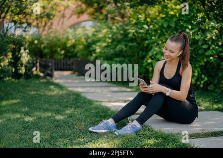 Felice atletica donna in sportsclothes sorride allegramente surfs telefono cellulare, mentre ascolta la musica tramite auricolari wireless prende pausa dopo il sole mornin Foto Stock