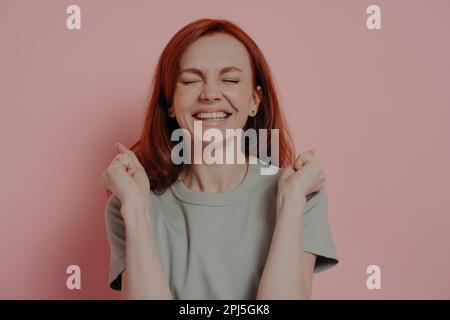 Primo piano scatto di felice femmina dai capelli rossi che fa desiderio con gli occhi chiusi, pugni aggrappati e sorridenti positivamente, vestito in abiti casual isola Foto Stock