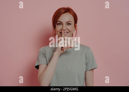 Giovane ragazza zenzero positivo fare un gesto di shush, tenendo l'indice sopra le labbra e chiedendo di non dire informazioni segrete, vestito con t-shirt casual, è Foto Stock