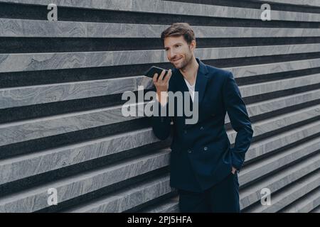 Un uomo adulto professionale e prospero con stoppia utilizza l'altoparlante per rispondere alle chiamate in arrivo lascia il messaggio audio vocale tiene il telefono cellulare moderno vicino alla bocca Foto Stock