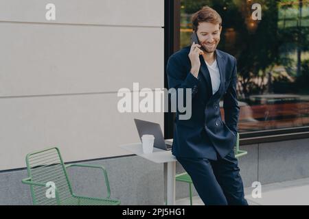 Buon commerciante maschio riuscito che discute i prezzi di azione sopra mentre si appoggia alla tabella del caffè del marciapiede, godendosi essere lavoratore autonomo, felice dopo aver sentito il goo Foto Stock