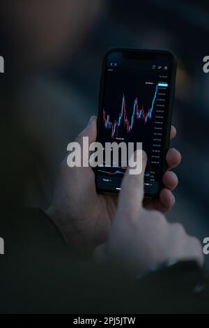 Trader che controlla i dati del mercato azionario nell'app mobile per il trading online. Uomo mano toccare lo schermo dello smartphone con grafico forex in tempo reale, lettura finanziaria Foto Stock