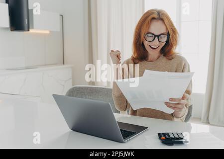 Il freelancer femminile di zenzero di successo controlla i calcoli clenches pugno guarda felicemente i documenti di carta indossa occhiali e jumper circondato da lapto Foto Stock