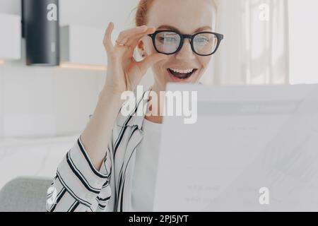 Positivo redhead giovane donna sorrisi indossa in generale occhiali trasparenti focalizzati su carte di controllo relazione finanziaria felice di finire il lavoro nel tempo opere da Foto Stock