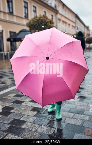 Bambina che si nasconde dietro un grande ombrello rosa che cammina in un centro in un giorno d'autunno piovoso e cupo Foto Stock
