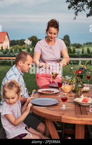 Famiglia che ha un pasto dalla griglia durante la cena estiva picnic all'aperto in un giardino di casa. Primo piano di persone sedute a un tavolo con cibo e piatti Foto Stock