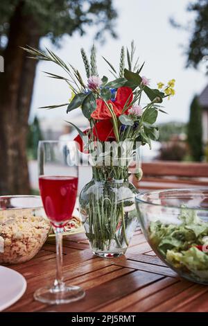 Cena in un giardino di meleti su un tavolo di legno con insalate e vino decorato con fiori. Primo piano del tavolo con il cibo preparato per la cena di famiglia Foto Stock