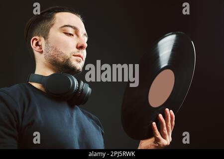 Uomo che guarda il disco in vinile. Stile musicale vintage. Maschio con cuffie che tengono il vecchio disco musicale in piedi su sfondo nero. Musica retrò. Audio classico Foto Stock