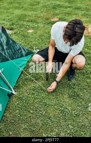 Giovane uomo che mette su una tenda in campeggio durante il viaggio estivo di vacanza. Teenager che mette i pali in terra erbosa usando il martello Foto Stock