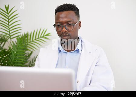 Occupato medico nero siede alla sua scrivania di lavoro sul suo computer portatile, guardando serio Foto Stock