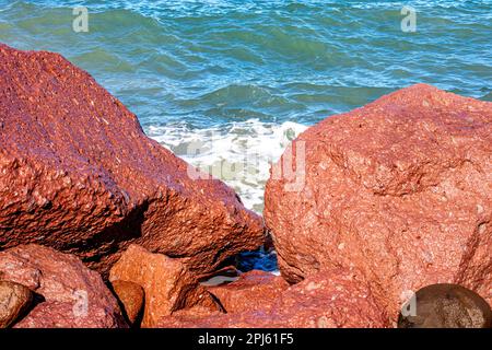 Rocce marroni rossastre bagnate con il mare e piccole onde sullo sfondo, costa rocciosa costiera, luce solare riflessa sull'acqua, giorno di sole a la Paz, Baj Foto Stock