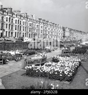 1950s, vista storica da questa epoca di persone che si trovano sulle panchine dei giardini sul lungomare di Brighton, Inghilterra, Regno Unito. La terrazza vittoriana era la casa di molti hotel che si occuperanno dei turisti. Un furgone dell'epoca è parcheggiato nella pubblicità di strada, Onchan Head... tutto il divertimento della fiera... visita la città bianca. Foto Stock