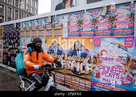 Street scene a Whitechapel, dove una guardia intorno a un cantiere edile è coperta da poster pubblicitari per varie serate di club il 8th marzo 2023 a Londra, Regno Unito. Foto Stock