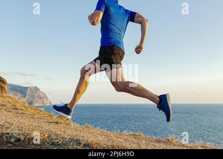 runner maschile in salita, sentiero sul mare su erba appassita, corsa di fondo, camicia blu e pantaloncini neri Foto Stock