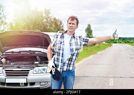 Uomo con strumenti in attesa di aiuto e che mostra i pollici fino nei pressi della sua vettura rotto Foto Stock