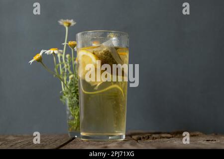 vetro trasparente con tè e fiori di camomilla su un tavolo di legno su grigio Foto Stock