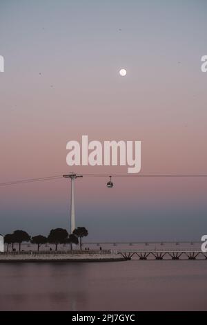 Una vista verticale e di basso profilo di una splendida scena caratterizzata da una luna piena, un tramonto dal cielo rosa, il fiume Tejo e una funivia. L'effetto complessivo è sereno Foto Stock