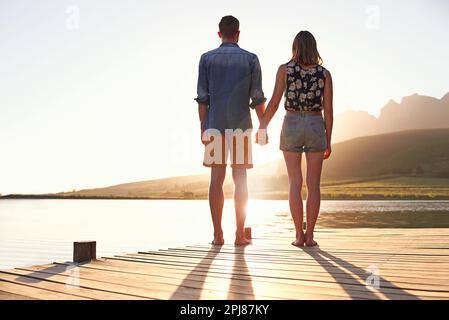 Prendere gli ultimi raggi di una giornata estiva. Ripresa da dietro di una giovane coppia affettuosa in piedi su un molo al tramonto. Foto Stock