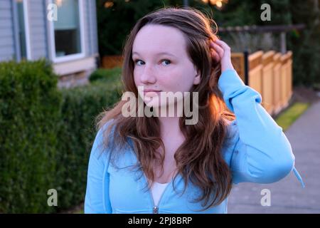 Bella giovane ragazza va e raddrizza hair.The tramonto illumina i capelli con retroilluminazione una blusa blu sulla strada contro lo sfondo della casa e cespugli giovane donna Foto Stock