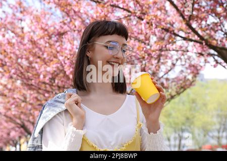 Bella giovane donna che tiene la tazza di caffè di carta nel parco con alberi sakura fiorenti Foto Stock