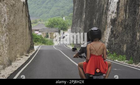 Una giovane coppia che indossa caschi su una motocicletta corre lungo la strada lungo le montagne rocciose. Un giovane uomo e una ragazza in un abito rosso estivo in un tropi Foto Stock