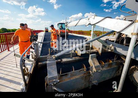 Vista sulla macchina per la posa di asfalto, stendendo strato di asfalto caldo su terreno preparato pochi lavoratori stanno lavorando intorno. Foto Stock