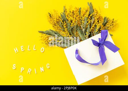 Fiori mimosa su borsa di carta regalo e l'iscrizione ciao primavera su sfondo giallo. Concetto di primavera, composizione dei fiori. Biglietto d'auguri. Visualizza da Foto Stock