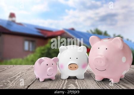 Piggy banche su superficie di legno e vista sfocata di bella casa. Concetto ipotecario Foto Stock