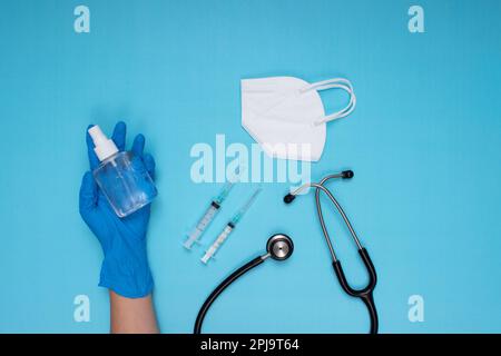 Guanti medici blu mano che tiene spray disinfettante, maschera chirurgica bianca, stetoscopio e aghi su sfondo azzurro Foto Stock