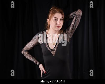 Giovane donna caucasica in piedi con un lieve sorriso, mano sull'anca e su un altro braccio sollevato, indossando un abito nero con maniche in lacy di fronte a sfondo nero Foto Stock