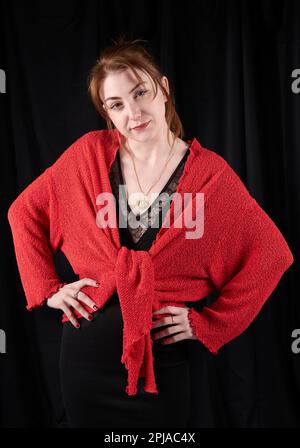 Giovane donna caucasica in piedi con un lieve sorriso, mani sull'anca, indossando cardigan rosso su abito nero di fronte a sfondo nero Foto Stock