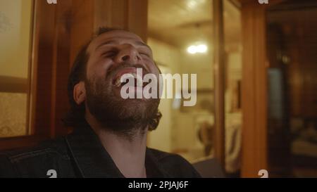 Giovane uomo in POV Holding Phone e laughing durante la videochiamata sul balcone di casa di legno illuminato di giallo Foto Stock