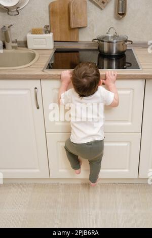 Il bambino salite su una stufa elettrica calda nella cucina domestica. Un bambino piccolo tocca la superficie della stufa con la mano al rischio di ottenere b Foto Stock