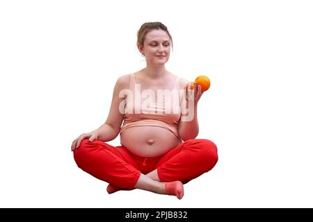 Una donna incinta si esercita a casa e tiene in mano un frutto come simbolo di cibo sano durante la gravidanza, isolato su uno sfondo bianco Foto Stock