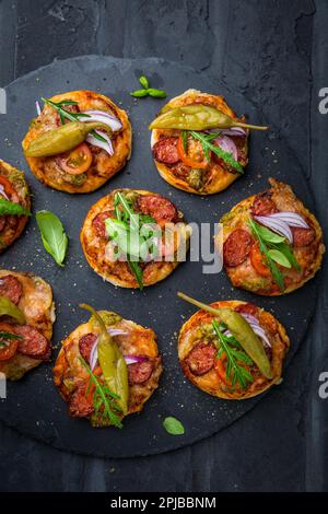 Piccola pizza fatta in casa con pesto, salame, pomodori e rucola su fondo nero Foto Stock