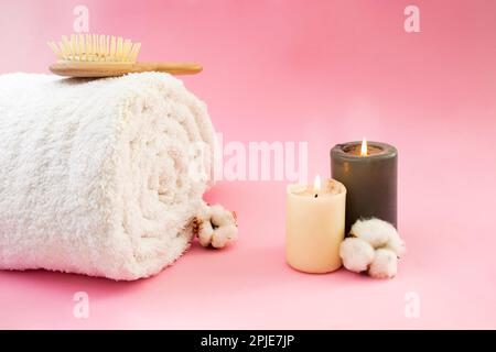 Asciugamano bianco arrotolato, spazzola per capelli di legno, fiori di cotone e candele accese sul dorso rosa Foto Stock