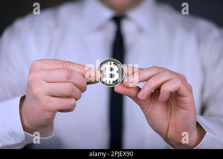 Un uomo d'affari in una camicia bianca e cravatta nera tiene una versione fisica di bitcoin nelle sue mani. Foto Stock