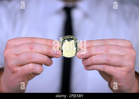 Un uomo d'affari in una camicia bianca e cravatta nera tiene una versione fisica di dogecoin nelle sue mani. Foto Stock