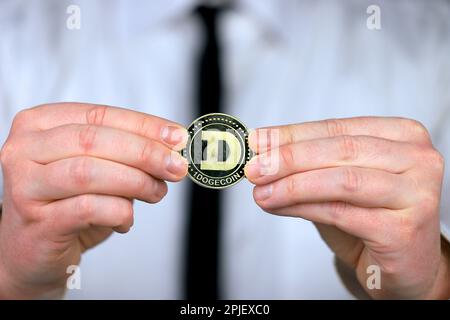 Un uomo d'affari in una camicia bianca e cravatta nera tiene una versione fisica di dogecoin nelle sue mani. Foto Stock
